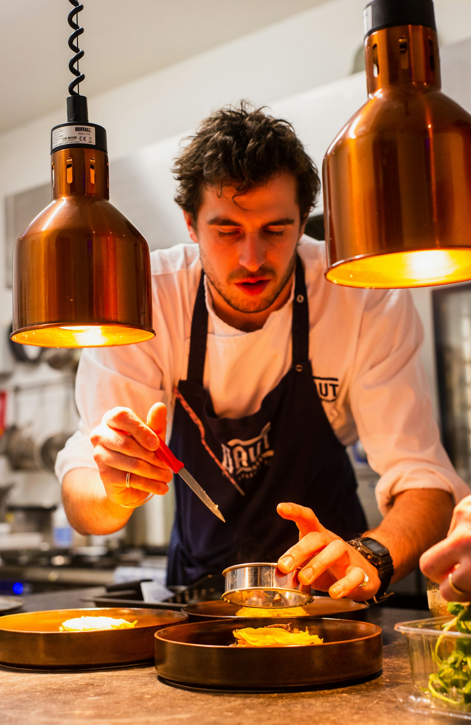 A chef plating a dish under copper heat lamps in a restaurant kitchen